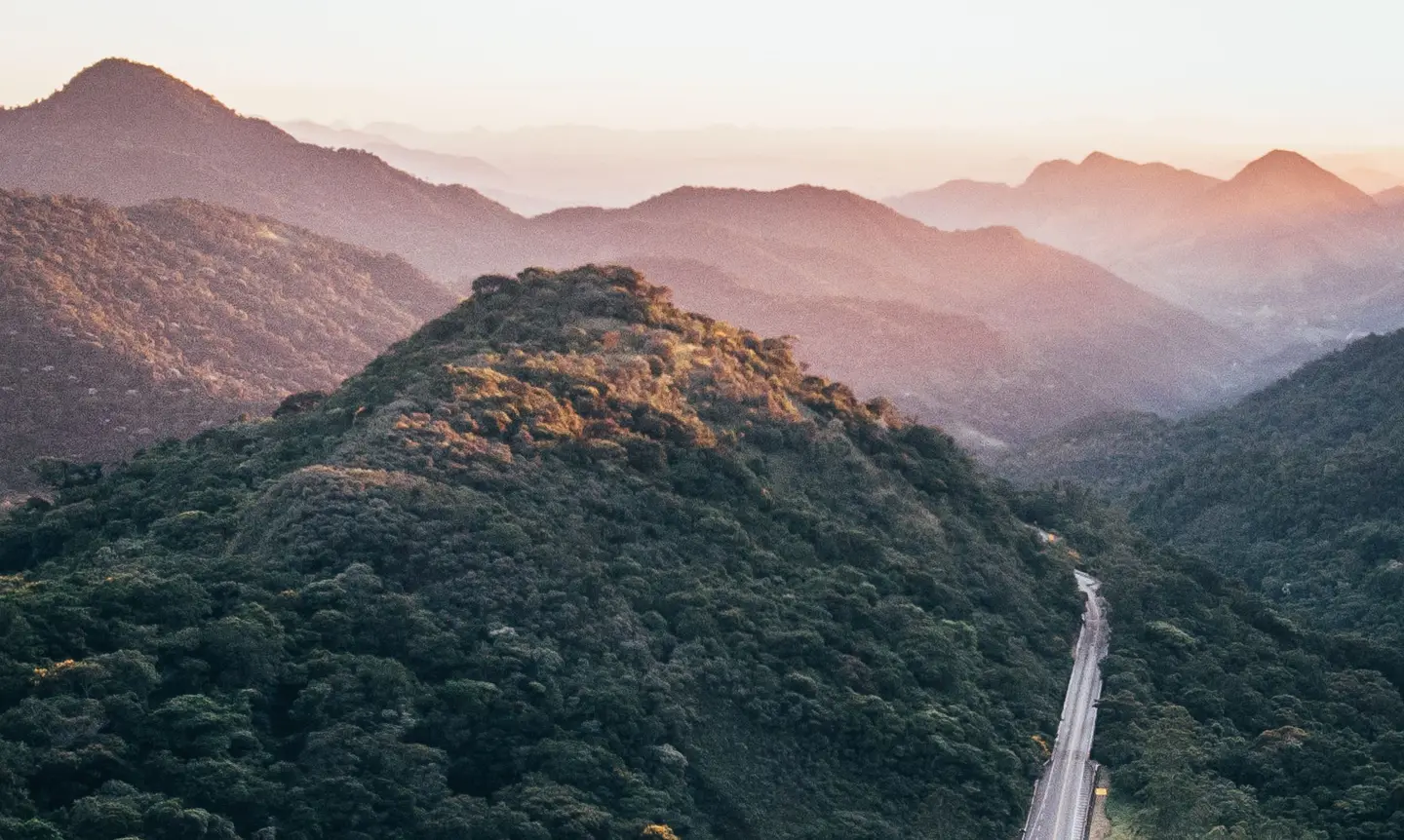 An aerial view at dusk of a highway curving through densely forested ridges that fade into haze on the horizon.